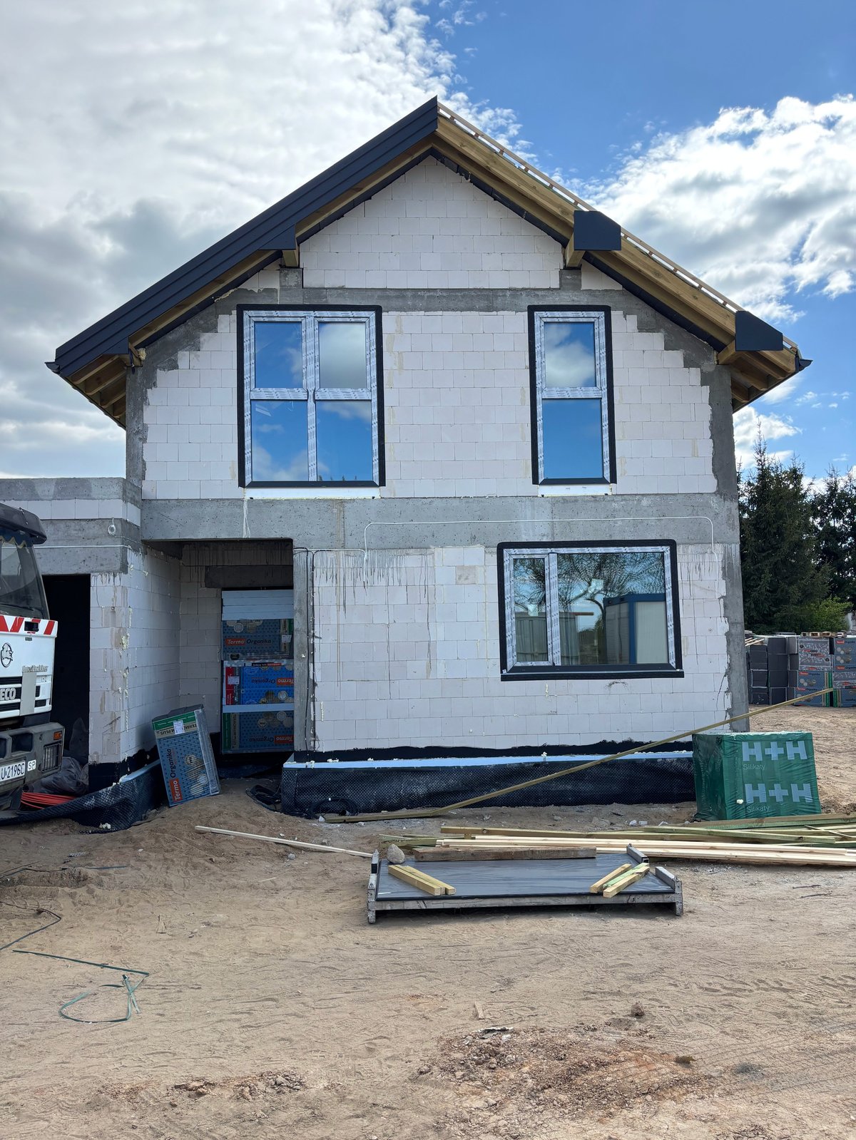 Two-story residential house under construction with white exterior, dark roof, and multiple windows surrounded by building materials and equipment on sandy ground