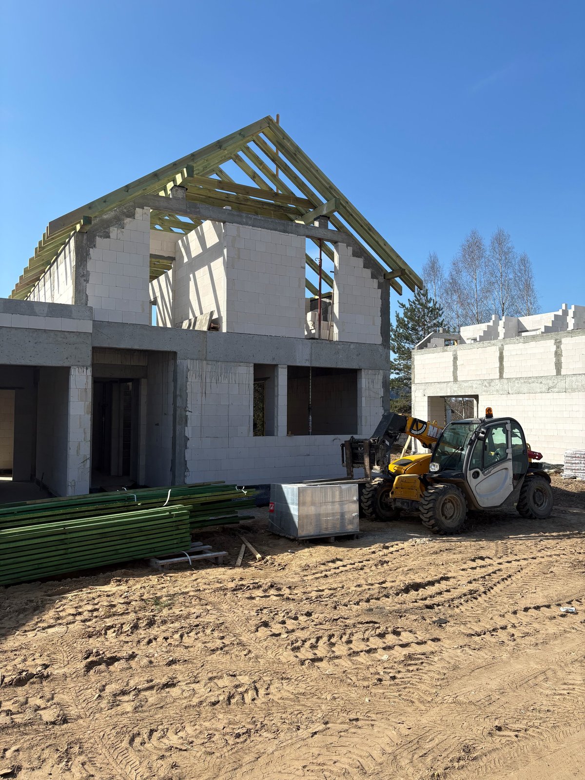 Construction site with a partially built concrete block house, wooden roof framing, and a yellow telescopic handler on sandy ground