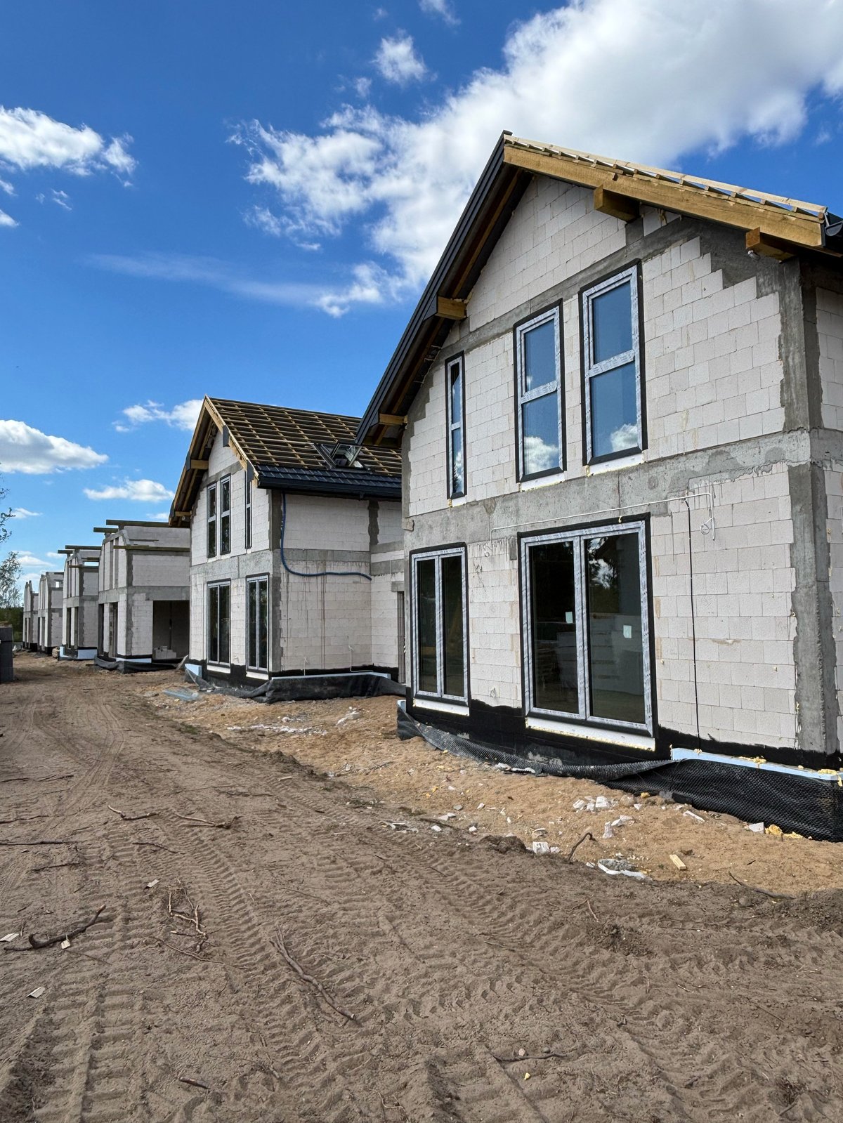 New residential houses under construction with white siding and large windows on unpaved lot under blue sky