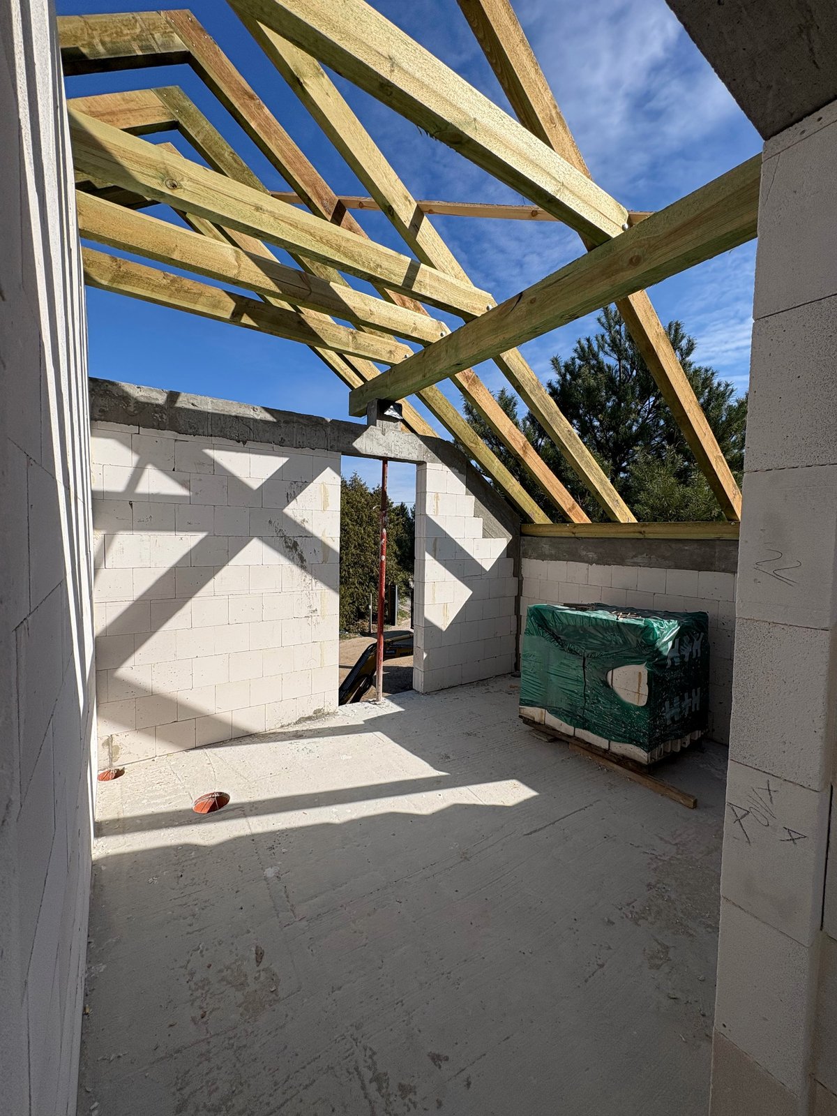 Under-construction patio with wooden pergola beams, concrete floor, white walls, and clear blue sky visible overhead