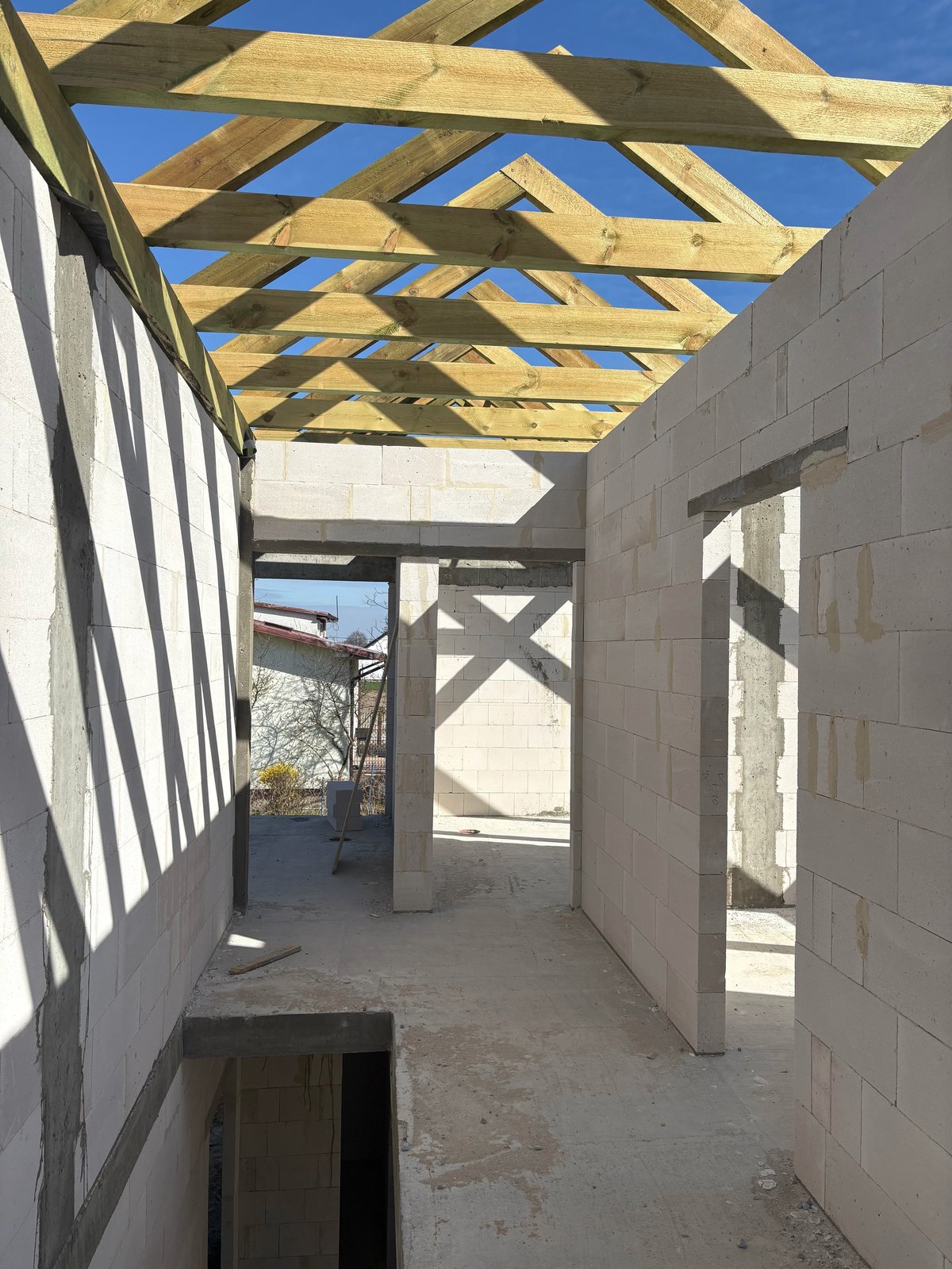 Interior of unfinished house with wooden beam ceiling framework, concrete block walls, and clear blue sky visible through open roof structure