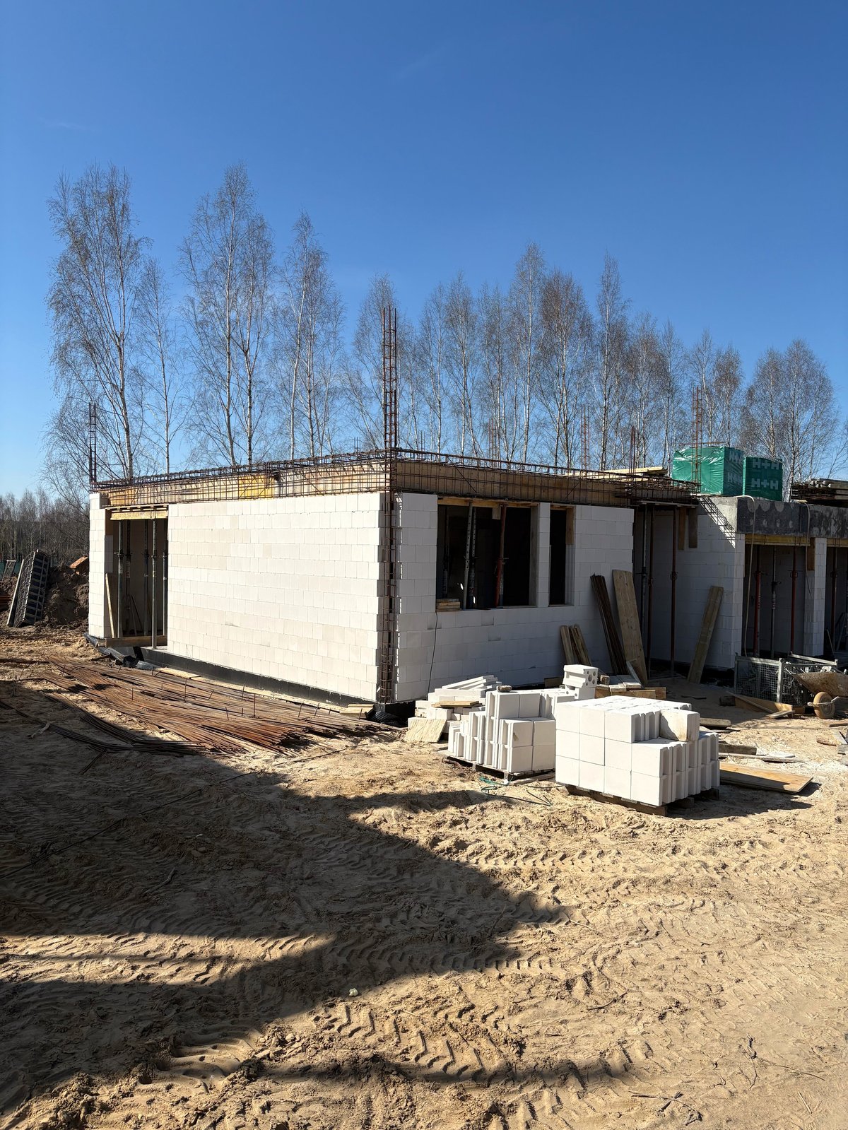 Construction site with a partially built residential structure, white concrete blocks, and birch trees under blue sky