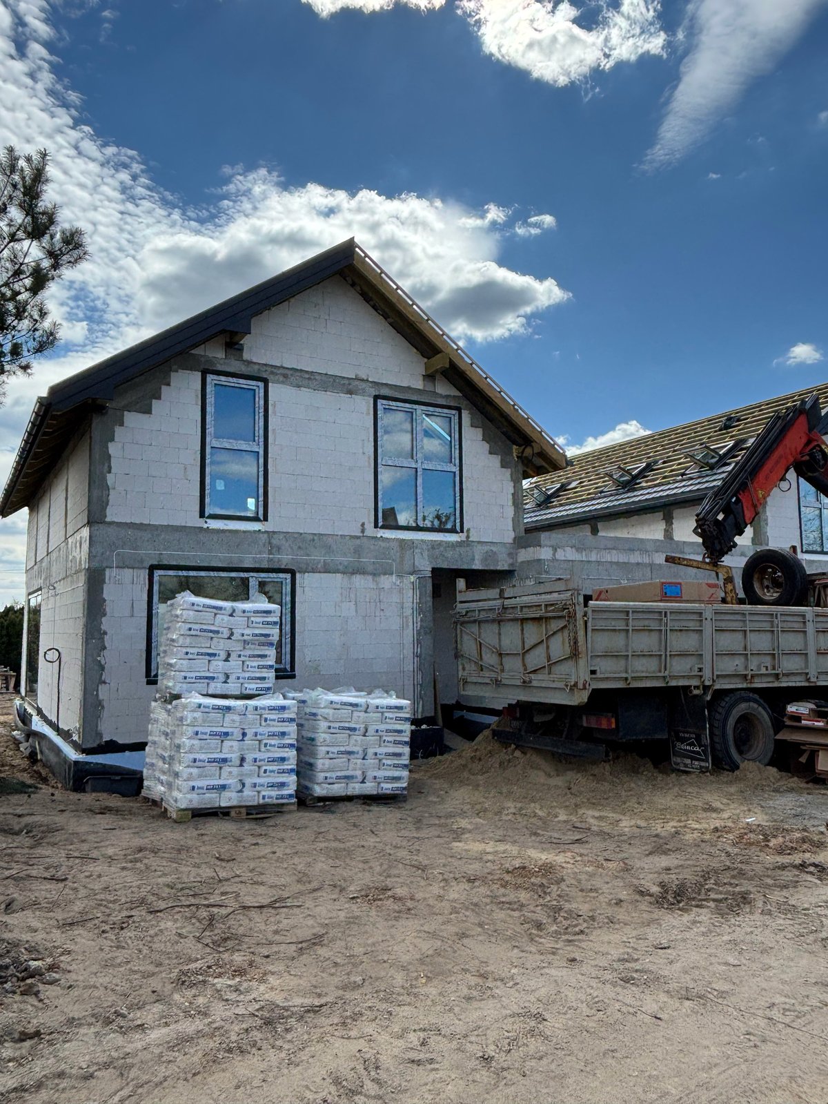 A two-story house under construction with white shingle siding, surrounded by building materials and a delivery truck