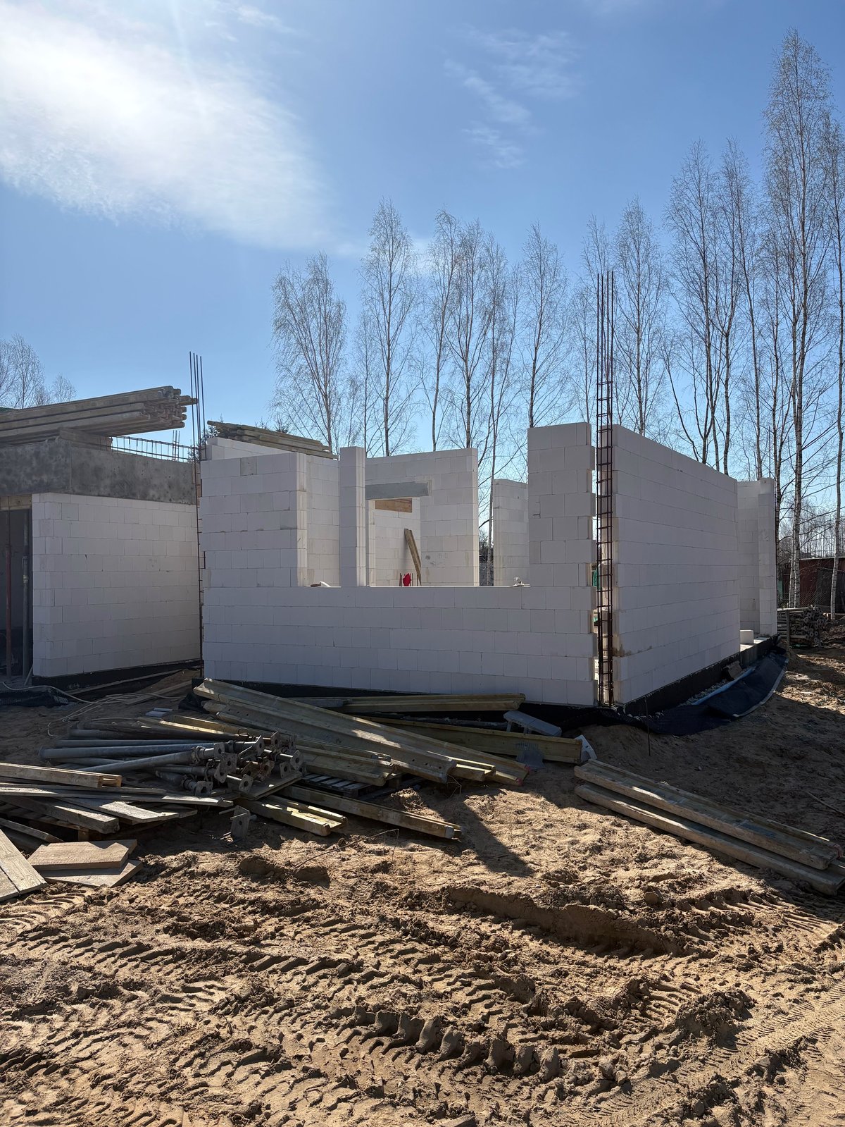 Unfinished white residential building under construction with bare birch trees in background and sandy ground in foreground