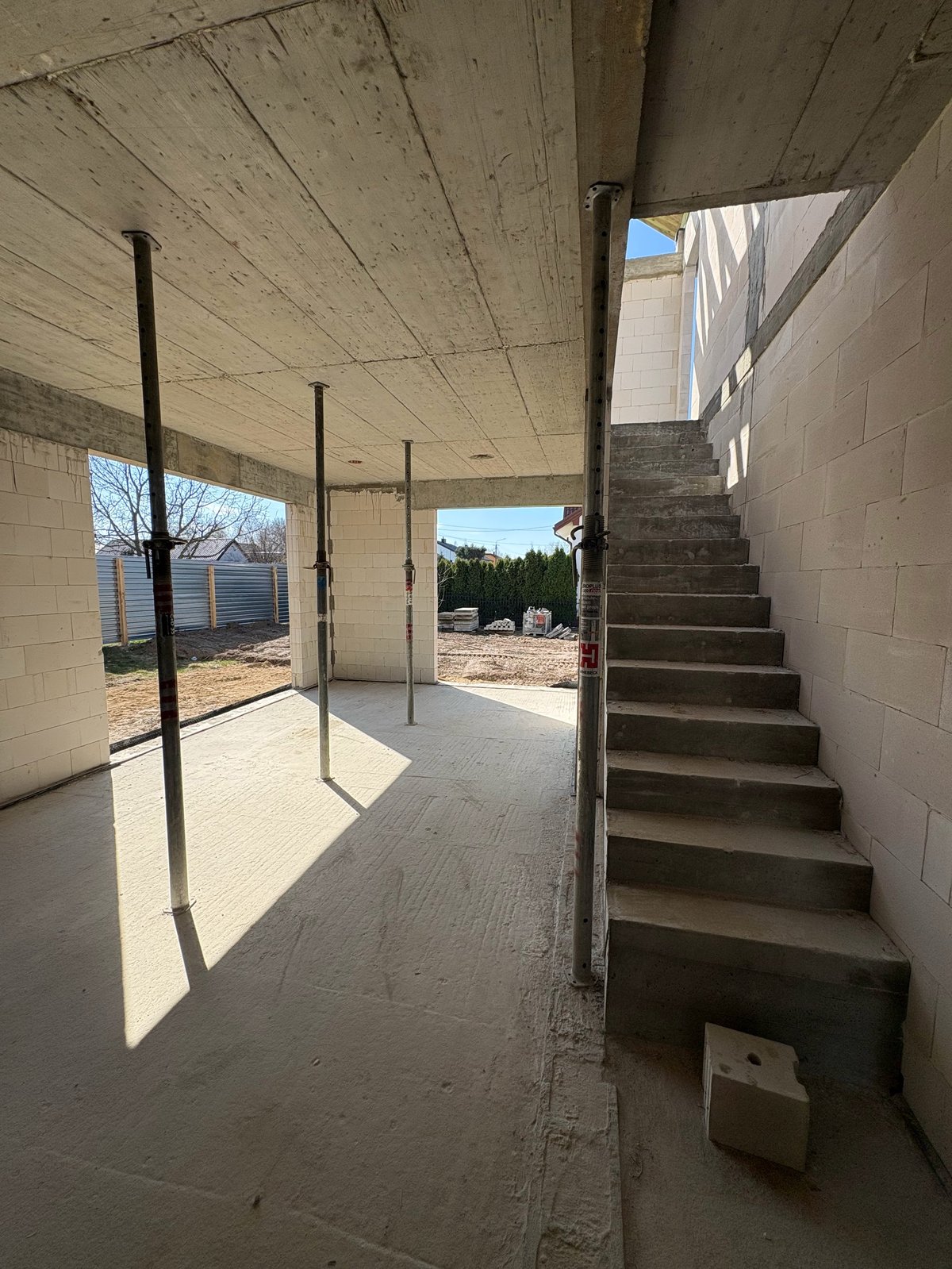 Interior of unfinished concrete basement with support posts, stairs, and bright sunlight streaming through openings
