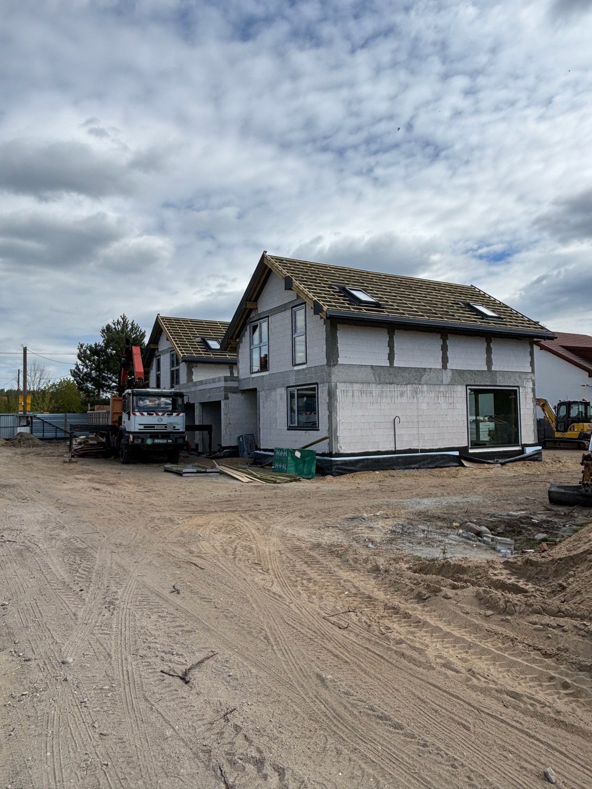 New house under construction with white walls and shingled roof on dirt lot surrounded by construction equipment and vehicles