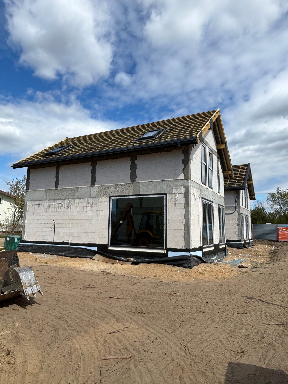New house under construction with white cladding, large windows, and shingled roof on dirt foundation under blue sky