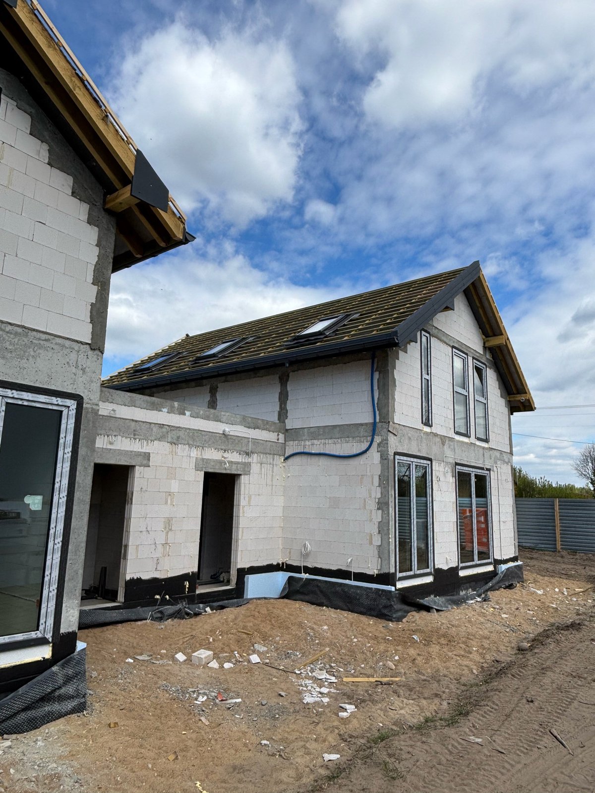 Modern house under construction with white brick exterior, pitched roof, and exposed foundation against blue sky